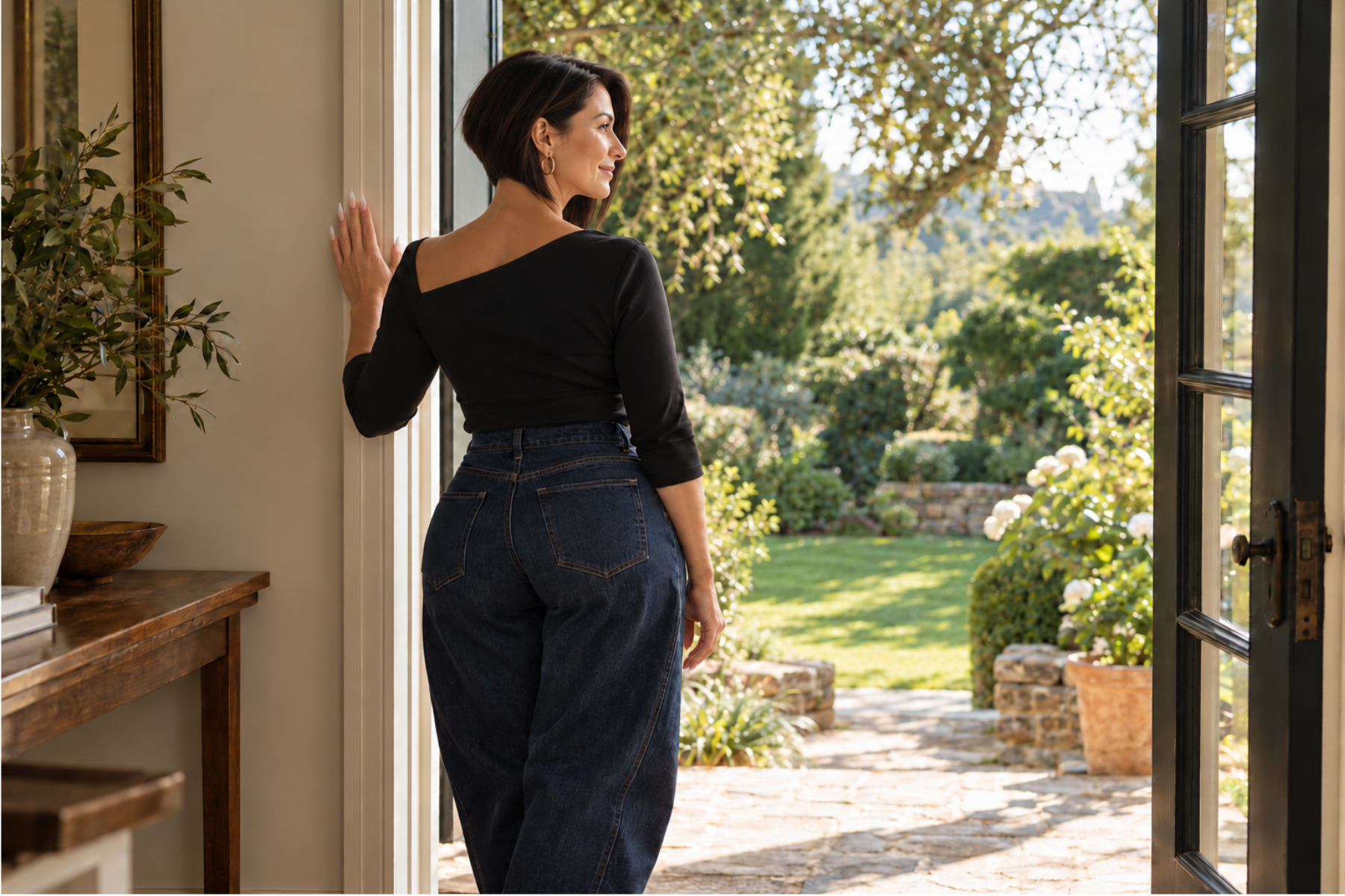 Woman with a sharp dark bob, black asymmetric top and dark denim jeans pauses at an open doorway overlooking a sunlit garden.